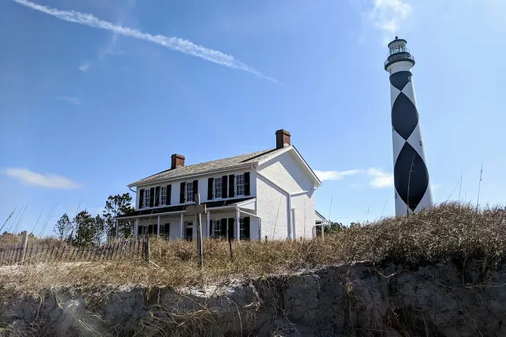 A white house and striped lighthouse on a sandy hill under a blue sky.